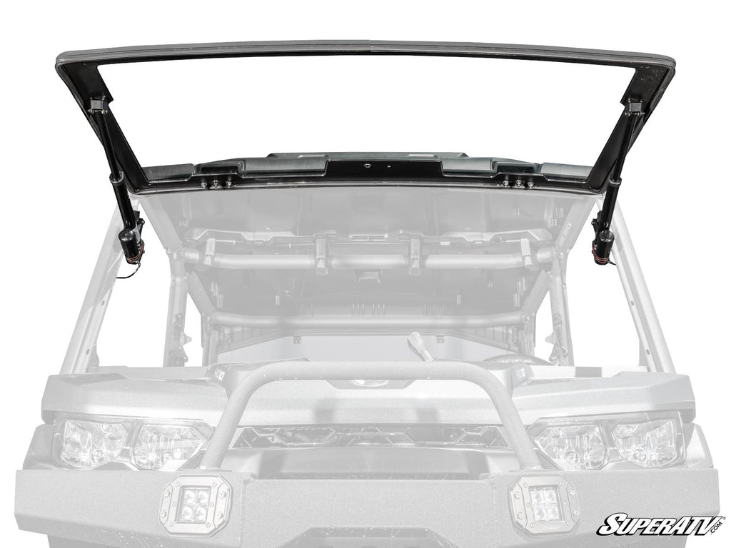 A grayscale image shows a Can-Am Defender with the MaxDrive Power Flip Glass Windshield, featuring a black, open flip-up design mounted in front. The SuperATV logo appears in the bottom right corner, emphasizing the windshield.