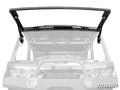 A grayscale image shows a Can-Am Defender with the MaxDrive Power Flip Glass Windshield, featuring a black, open flip-up design mounted in front. The SuperATV logo appears in the bottom right corner, emphasizing the windshield.