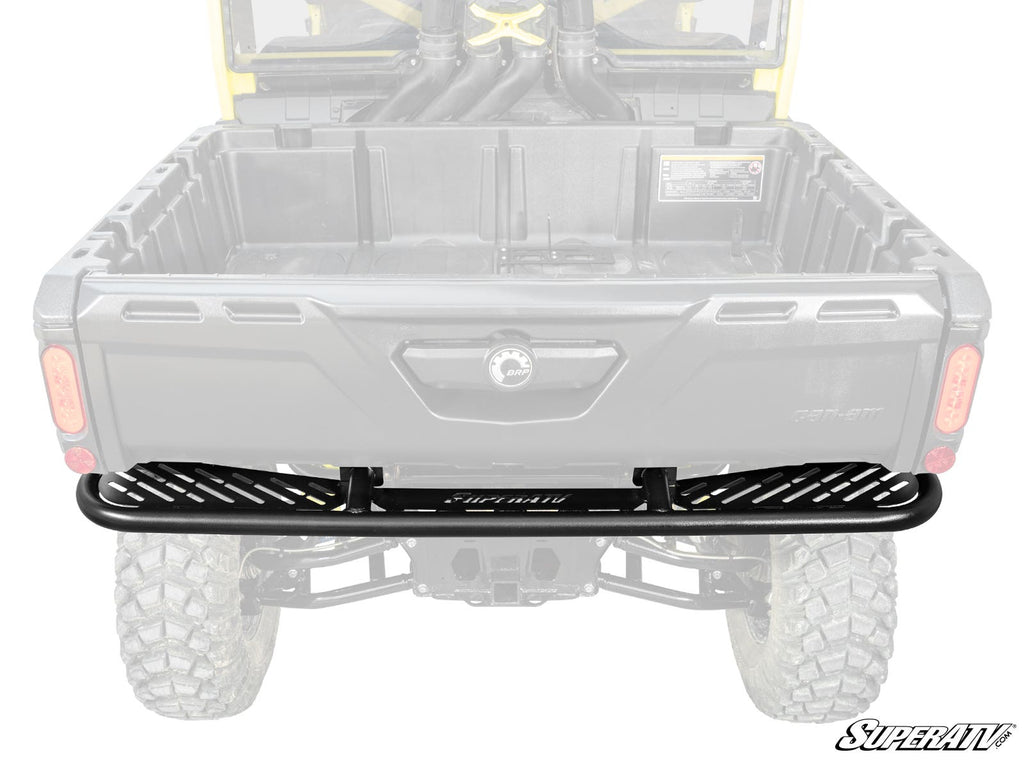 The Can-Am Defender Rear Bumper, made of black metal with a slotted design and SuperATV logo centered, is installed on the back of a Can-Am Defender. The bumper stands out prominently against the faded vehicle in the background.