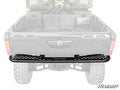 The Can-Am Defender Rear Bumper, made of black metal with a slotted design and SuperATV logo centered, is installed on the back of a Can-Am Defender. The bumper stands out prominently against the faded vehicle in the background.