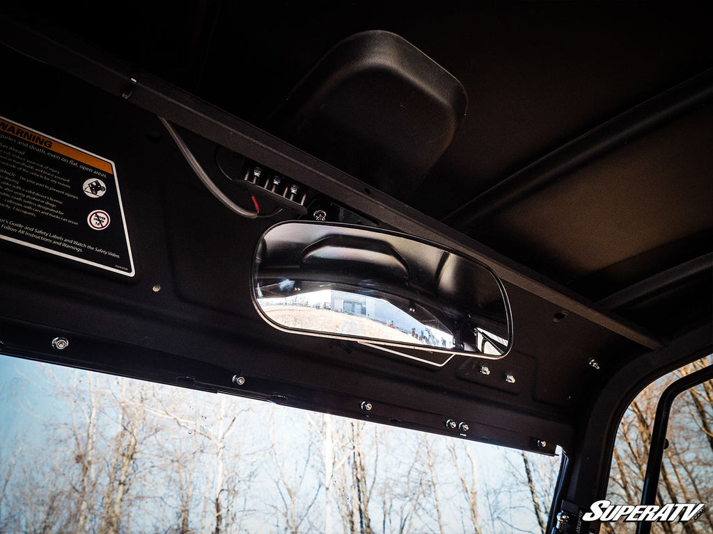 The interior of a Can-Am Defender shows the Rear View Mirror by SuperATV mounted above the dashboard, reflecting an outdoor scene with blue sky and leafless trees. The SuperATV logo is at bottom right; warning labels are visible inside.