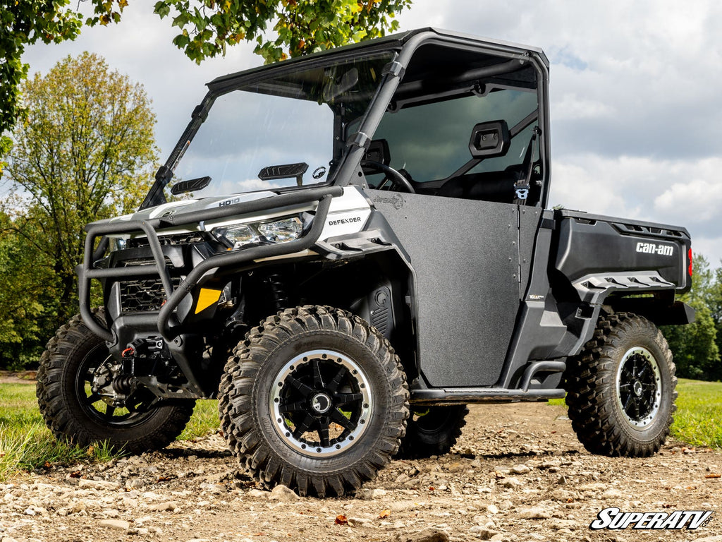 A rugged Can-Am Defender, equipped with a Front 2 Receiver Hitch, sits outdoors on a dirt path. It features large off-road tires, a front bumper guard, clear doors, and displays the SuperATV logo in the lower right corner.