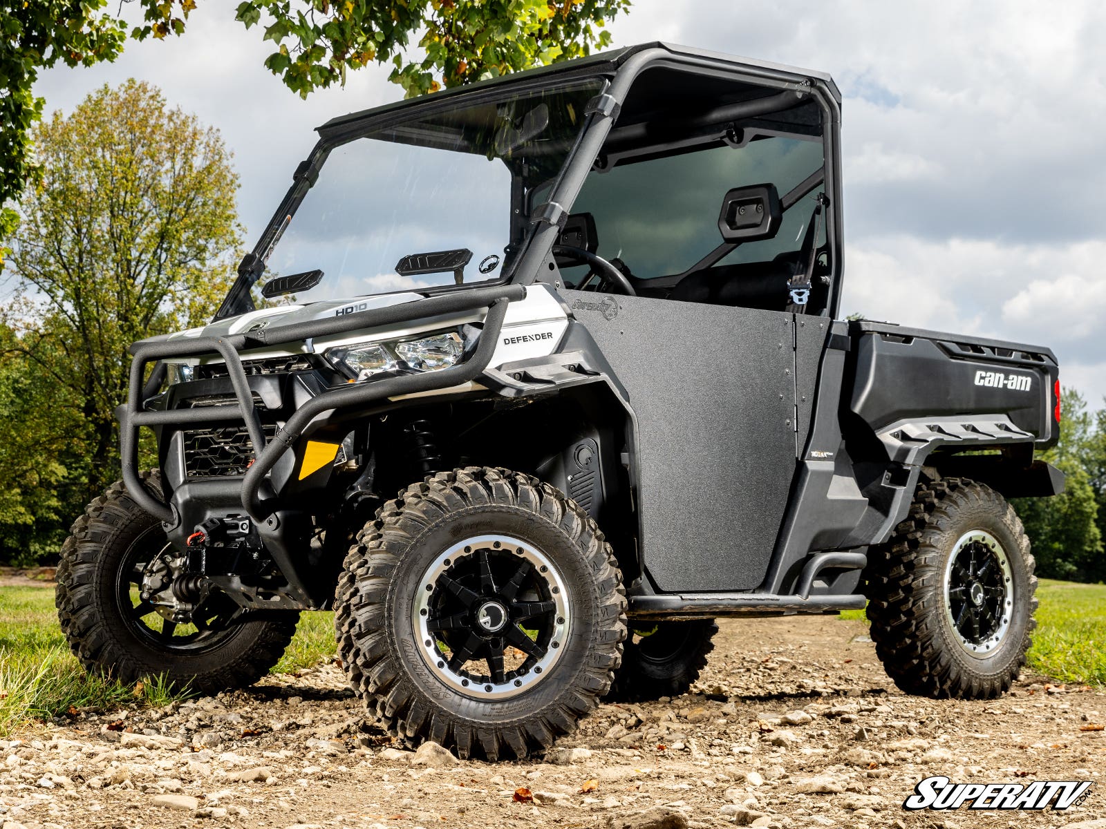 A rugged Can-Am Defender, equipped with a Front 2 Receiver Hitch, sits outdoors on a dirt path. It features large off-road tires, a front bumper guard, clear doors, and displays the SuperATV logo in the lower right corner.