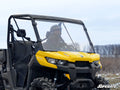 A rider in safety gear drives off-road in a bright yellow Can-Am HD10 equipped with the Can-Am Defender Full Windshield. Rugged tires and bare trees are visible outdoors under a cloudy sky, with the SuperATV logo shown in the corner.