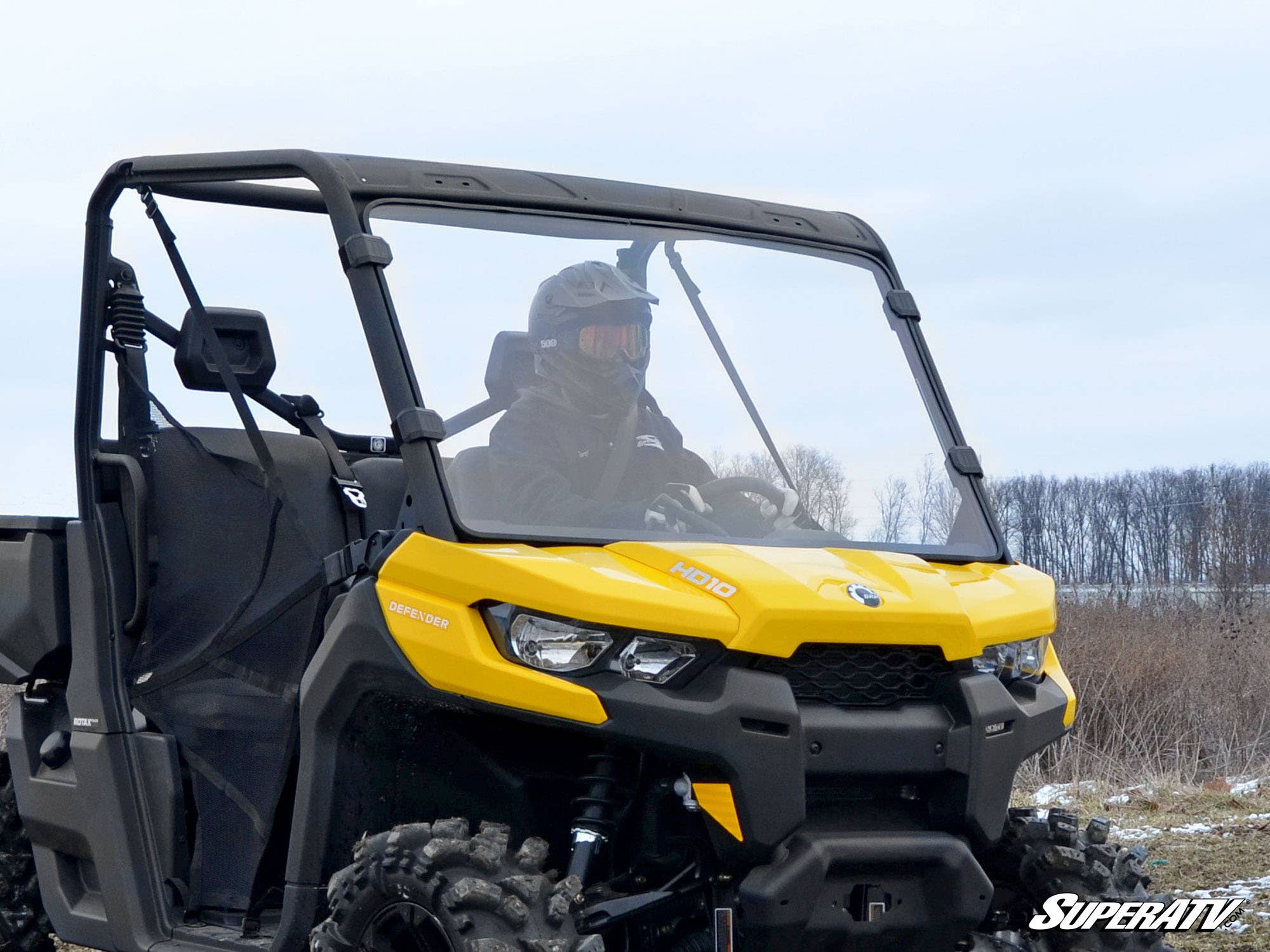 A rider in safety gear drives off-road in a bright yellow Can-Am HD10 equipped with the Can-Am Defender Full Windshield. Rugged tires and bare trees are visible outdoors under a cloudy sky, with the SuperATV logo shown in the corner.
