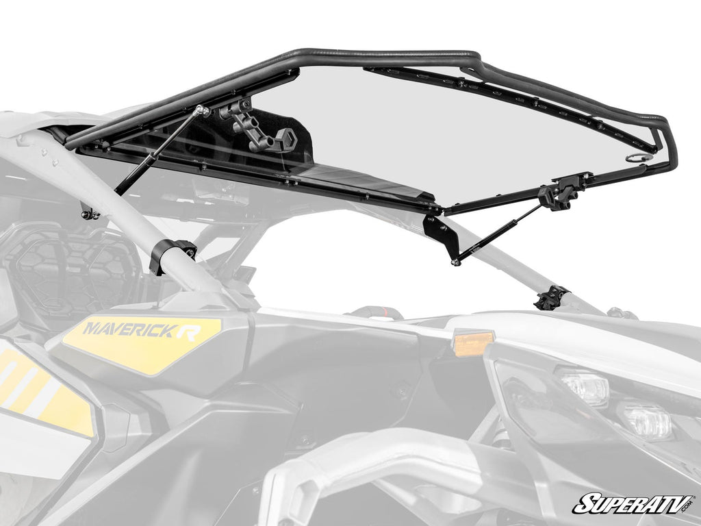 A clear, hinged Can-Am Maverick R Scratch-Resistant Flip Windshield is attached to the front of a white and yellow off-road vehicle. The partially open windshield is supported by metal arms, with the SuperATV logo in the lower right corner.