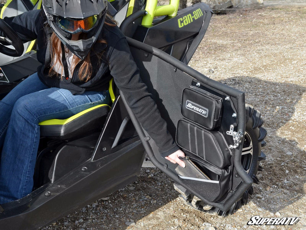 A person in a black helmet and jacket opens a mesh door on a black and green Can-Am off-road vehicle equipped with Can-Am Maverick Door Bags - Set of 2. The SuperATV logo appears at the bottom right, and the ground is rocky.