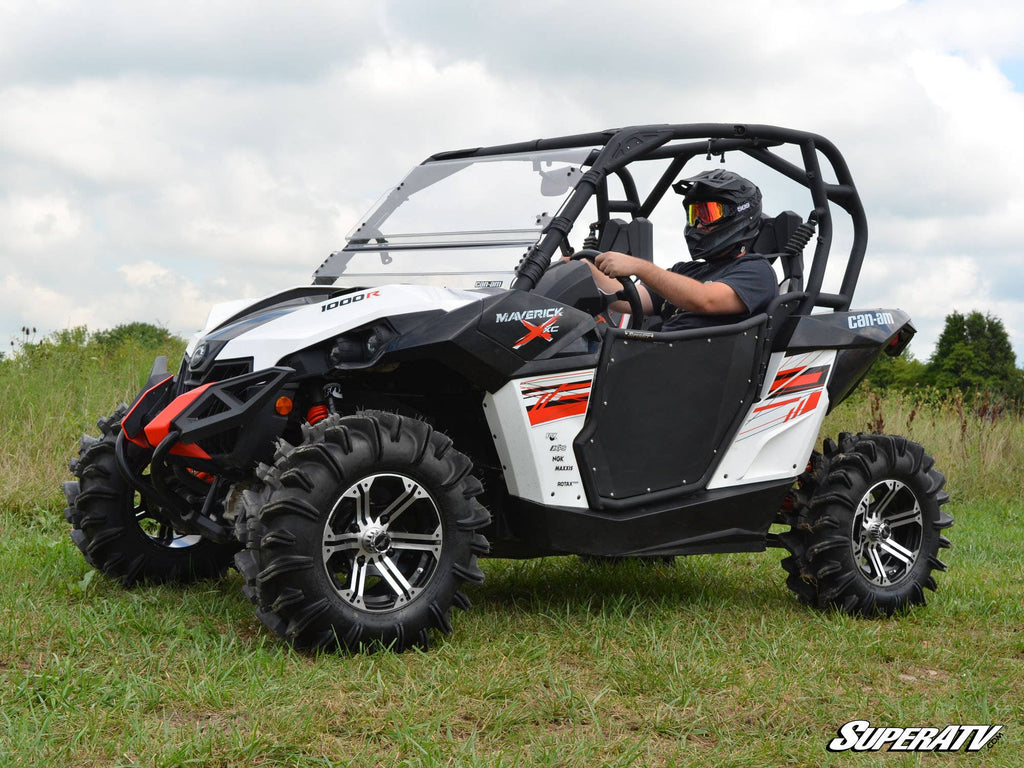 A person in a helmet drives a white and black Can-Am Maverick 1000R with Can-Am Maverick Aluminum Doors on grassy terrain. Rugged tires, red graphics, and the SuperATV logo appear; the sky is partly cloudy.