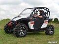 A person in a helmet drives a white and black Can-Am Maverick 1000R with Can-Am Maverick Aluminum Doors on grassy terrain. Rugged tires, red graphics, and the SuperATV logo appear; the sky is partly cloudy.