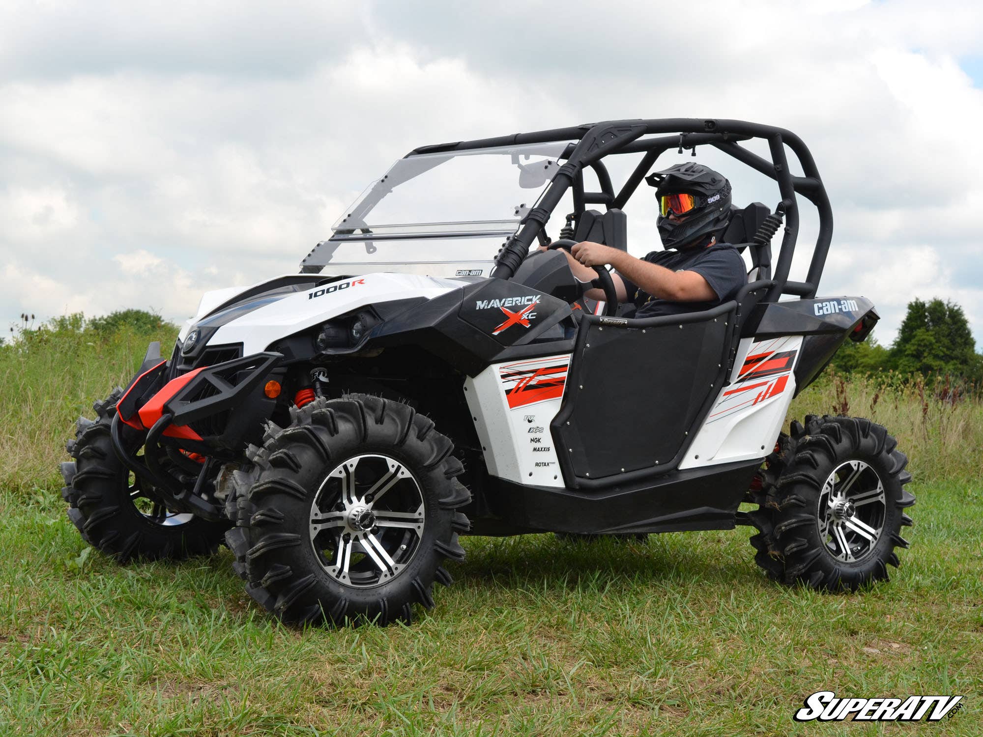 A person in a helmet drives a white and black Can-Am Maverick 1000R with Can-Am Maverick Aluminum Doors on grassy terrain. Rugged tires, red graphics, and the SuperATV logo appear; the sky is partly cloudy.