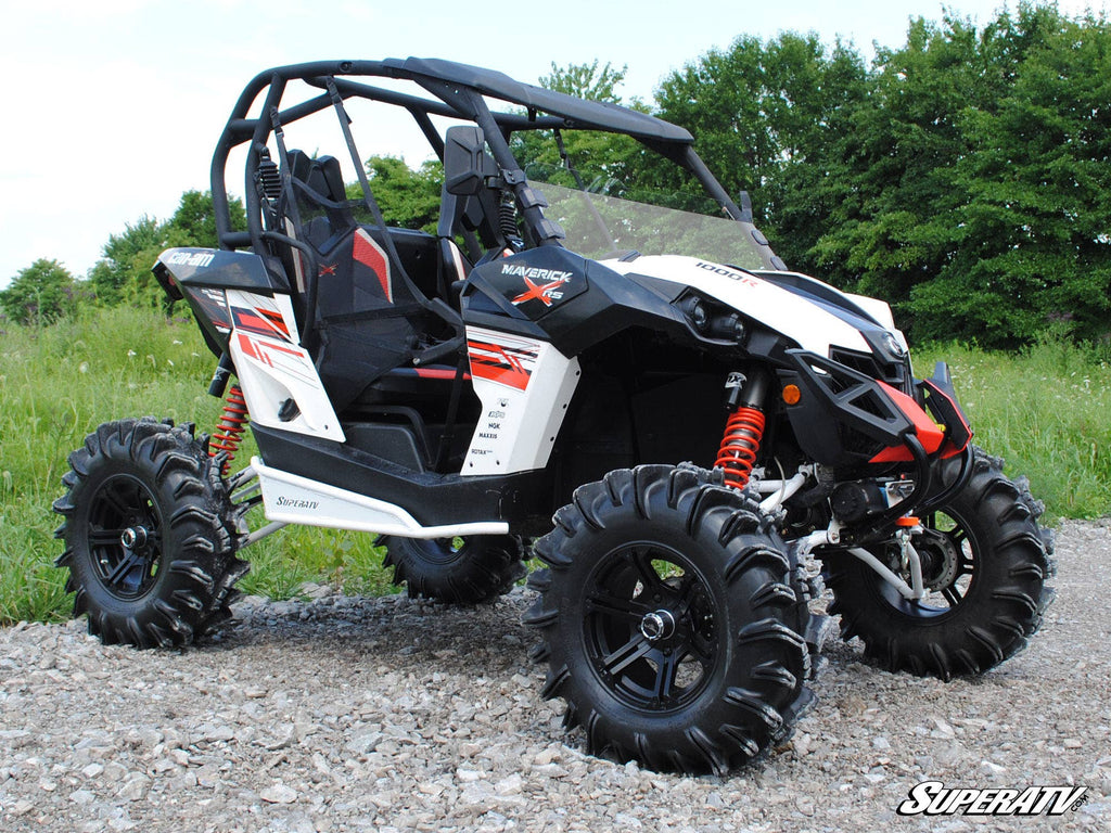 A Can-Am Maverick with a half windshield, roll cage, red and black decals, oversized deep-tread tires, and red suspension springs sits on a rocky path surrounded by green grass and trees under a clear sky.