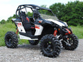 A Can-Am Maverick with a half windshield, roll cage, red and black decals, oversized deep-tread tires, and red suspension springs sits on a rocky path surrounded by green grass and trees under a clear sky.