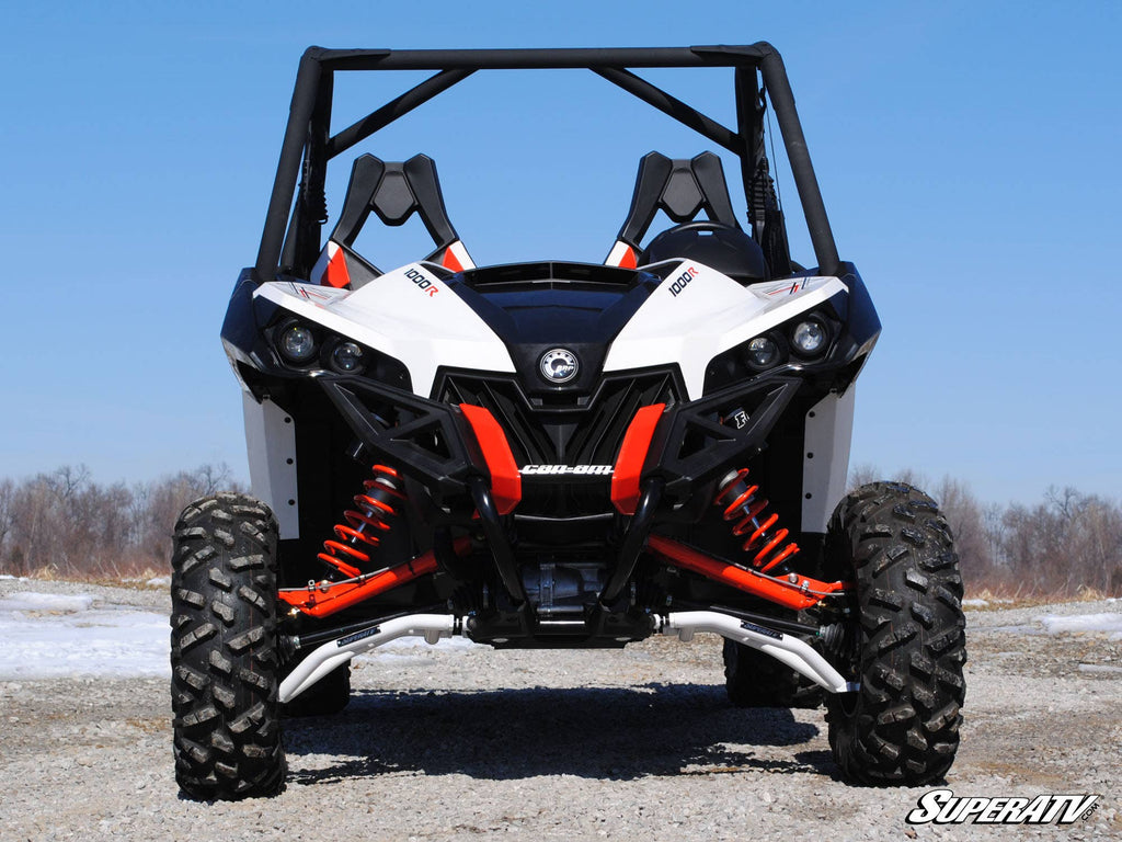 A front view shows a Can-Am off-road side-by-side with Maverick High Clearance Front A-Arms, rugged tires, orange suspension springs, bucket seats, and a roll cage on a gravel path amid a wintry landscape with bare trees.