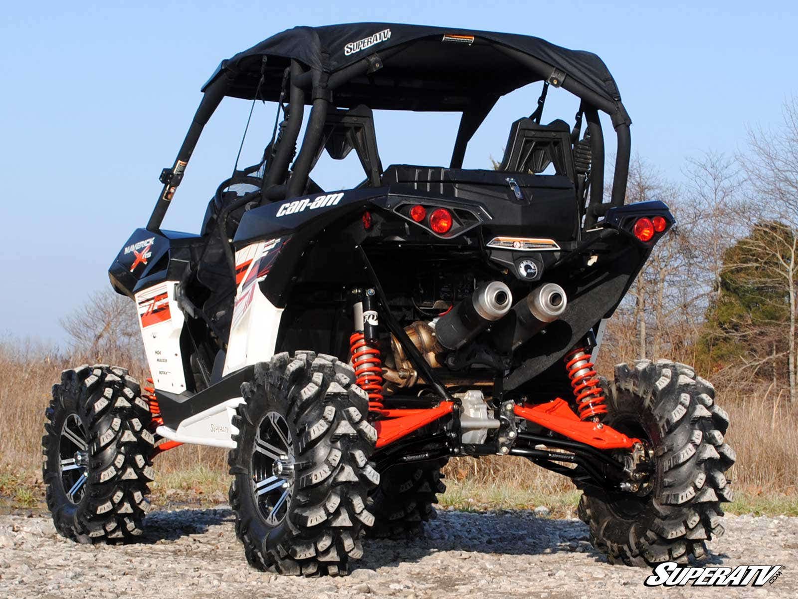 A Can-Am Maverick featuring High Clearance Rear A-Arms and orange suspension is parked on gravel outdoors. The black and white side-by-side has large rugged tires, with the SuperATV logo visible, set against bare trees and dry grass.