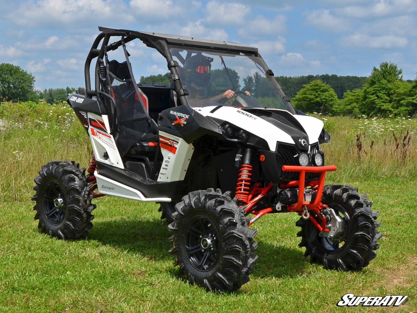 A Can-Am Maverick with a 4 Portal Gear Lift, featuring black and white bodywork, red suspension springs, and rugged tires, is parked on grass. It has two seats, an open frame, windshield, and a helmeted driver. Blue sky and trees are visible.