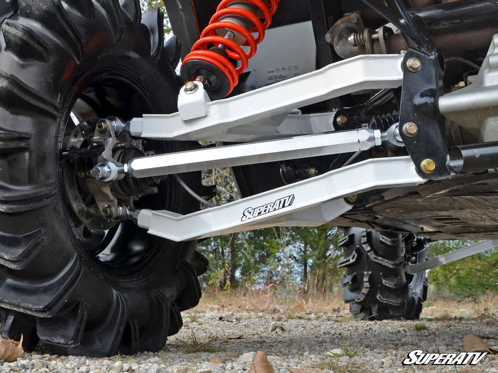 Close-up of a Can-Am Maverick’s suspension system featuring large black treaded tires, silver Can-Am Maverick Billet Aluminum Radius Arms labeled “SuperATV,” and a red coil spring, on rocky grass with trees in the background.