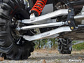 Close-up of a Can-Am Maverick’s suspension system featuring large black treaded tires, silver Can-Am Maverick Billet Aluminum Radius Arms labeled “SuperATV,” and a red coil spring, on rocky grass with trees in the background.