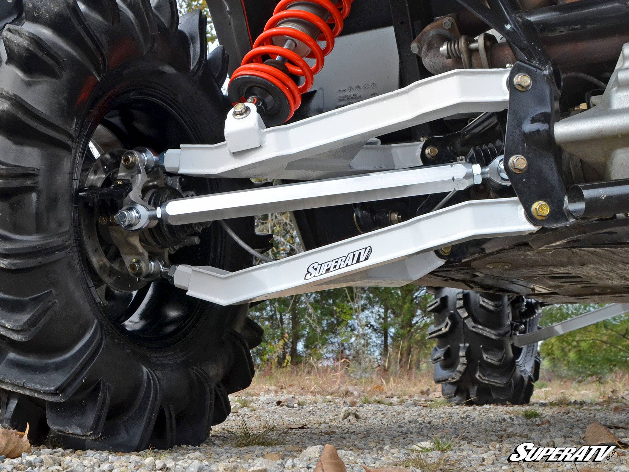 Close-up of a Can-Am Maverick’s suspension system featuring large black treaded tires, silver Can-Am Maverick Billet Aluminum Radius Arms labeled “SuperATV,” and a red coil spring, on rocky grass with trees in the background.
