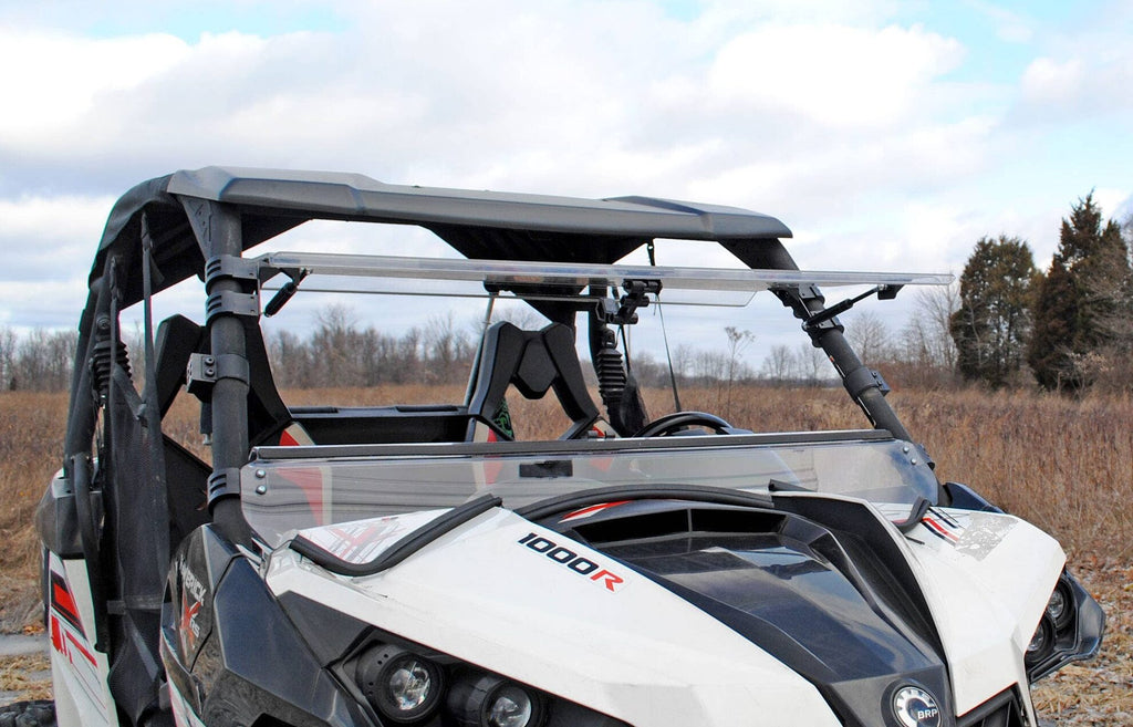A white side-by-side with a Can-Am Maverick Flip Windshield and flat roof is parked on grass under partly cloudy skies, with 1000R on its front and leafless trees in the background.
