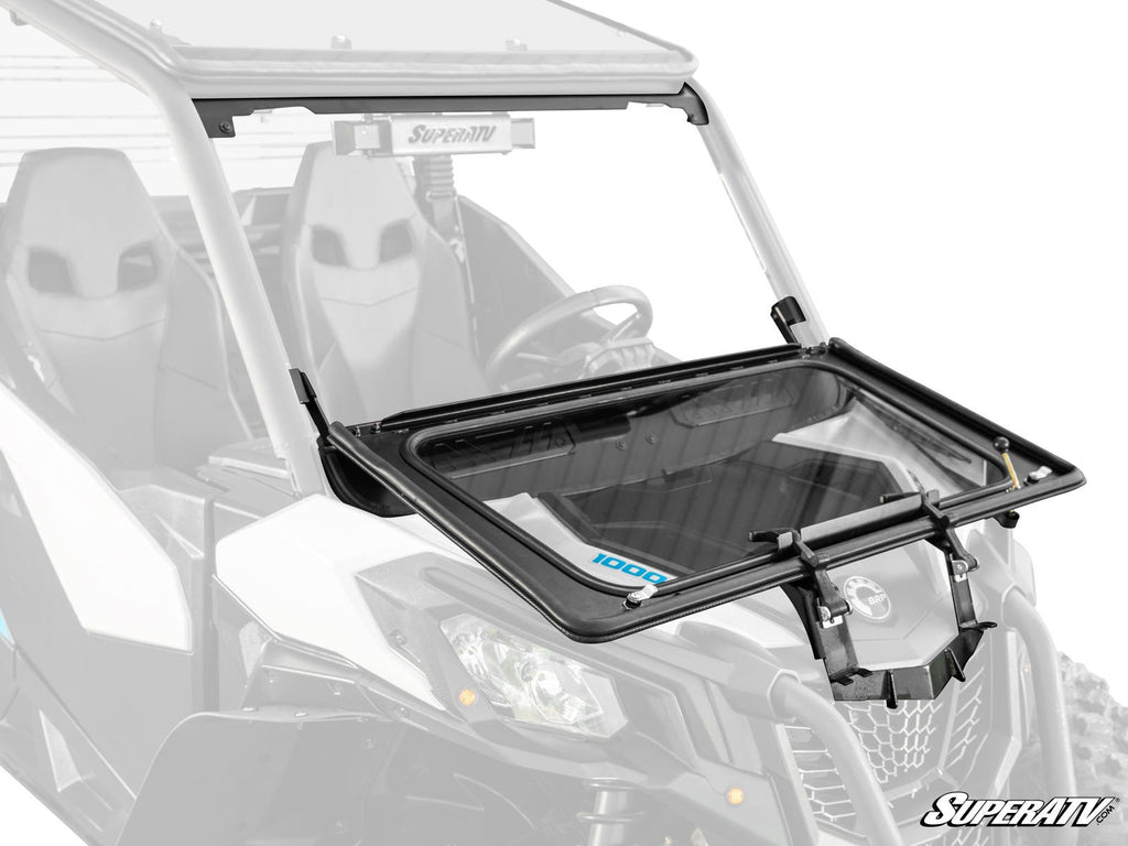 A close-up of a UTV displays the Can-Am Maverick Sport Flip Down Glass Windshield, partially opened to show its black frame and tilting hinges. The SuperATV logo is in the bottom right. Two harnessed seats are visible inside.