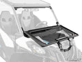 A close-up of a UTV displays the Can-Am Maverick Sport Flip Down Glass Windshield, partially opened to show its black frame and tilting hinges. The SuperATV logo is in the bottom right. Two harnessed seats are visible inside.