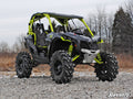 A Can-Am Maverick Turbo with a 3 Lift Kit, featuring large rugged tires, roll cage, exposed suspension, and racing seats in black and neon yellow, is parked on rocky ground with leafless trees and dry grass beneath a cloudy sky.