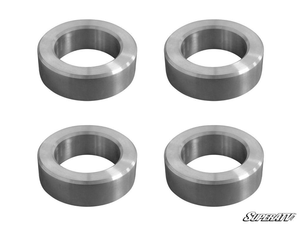 Four identical metal spacers with hollow centers are arranged in a 2x2 grid on a white background, each featuring a smooth, shiny finish. The SuperATV logo is visible in the bottom right corner. Product shown: Can-Am Maverick X3 3 Lift Kit.