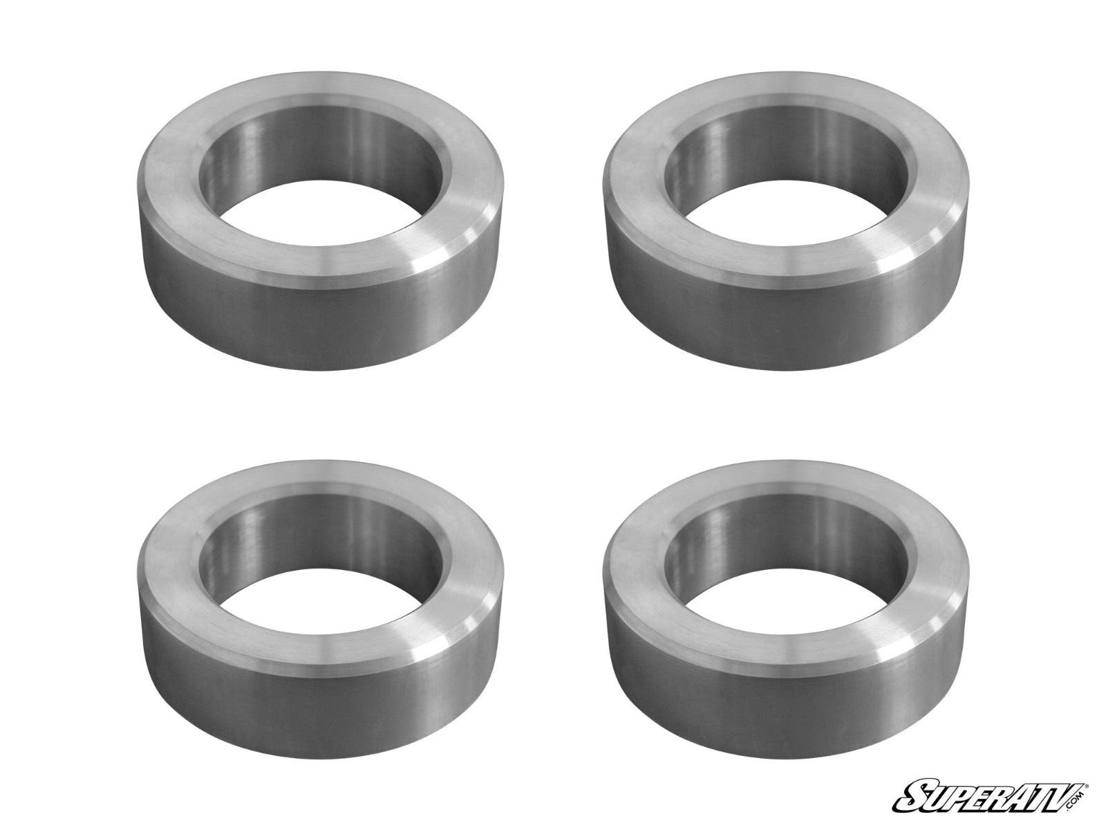 Four identical metal spacers with hollow centers are arranged in a 2x2 grid on a white background, each featuring a smooth, shiny finish. The SuperATV logo is visible in the bottom right corner. Product shown: Can-Am Maverick X3 3 Lift Kit.