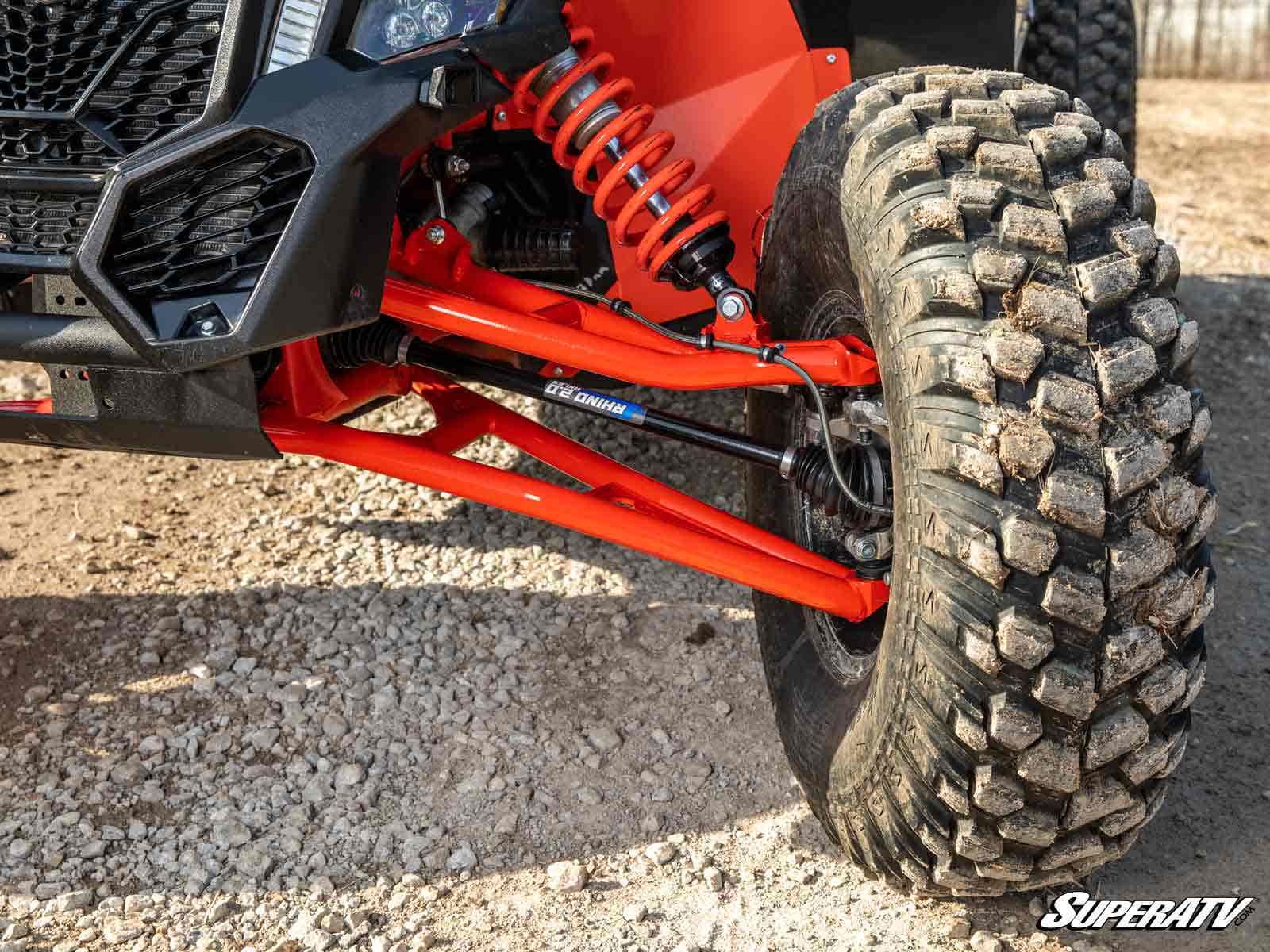 Close-up of a Can-Am Maverick X3 Atlas Pro 2 Forward Offset A-Arms with bright red control arms, large coilover shock, and mud-terrain tire. Gravelly ground and the SuperATV logo appear in the lower right corner.