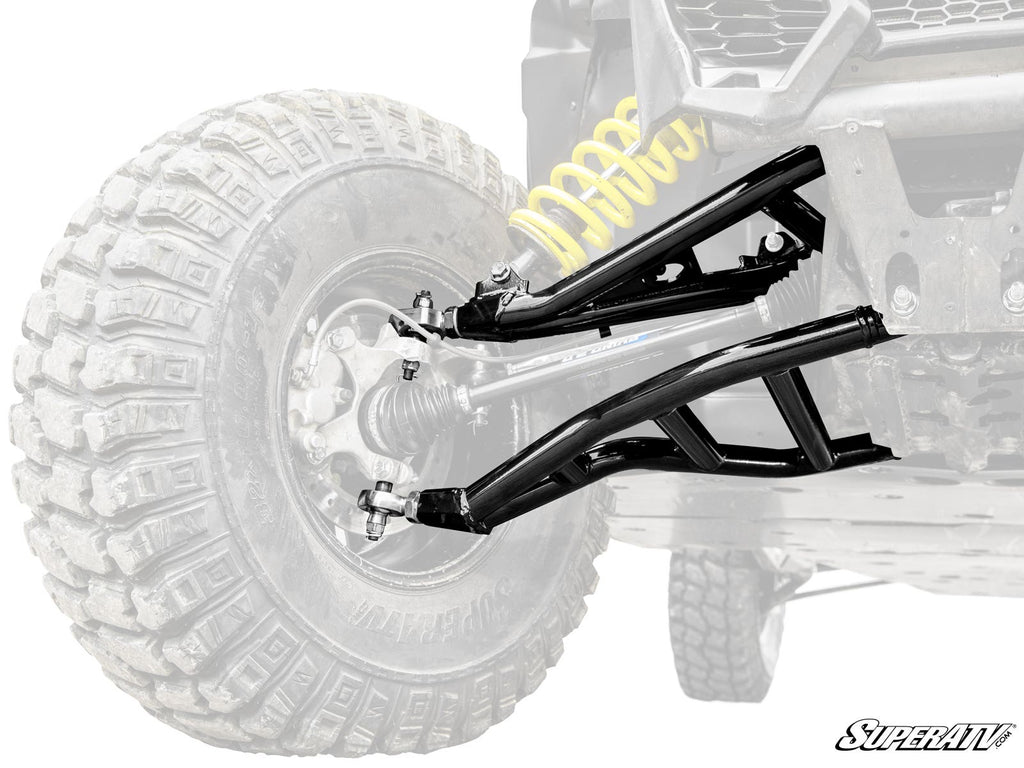 Close-up of Can-Am Maverick X3 Atlas Pro Heim Joint 1.5 Forward Offset A-Arms in shiny black, attached to a rugged off-road vehicle with large knobby tires; yellow coil spring stands out. “SuperATV” logo appears bottom right. Background vehicle is faded f