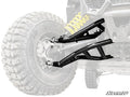 Close-up of Can-Am Maverick X3 Atlas Pro Heim Joint 1.5 Forward Offset A-Arms in shiny black, attached to a rugged off-road vehicle with large knobby tires; yellow coil spring stands out. “SuperATV” logo appears bottom right. Background vehicle is faded f