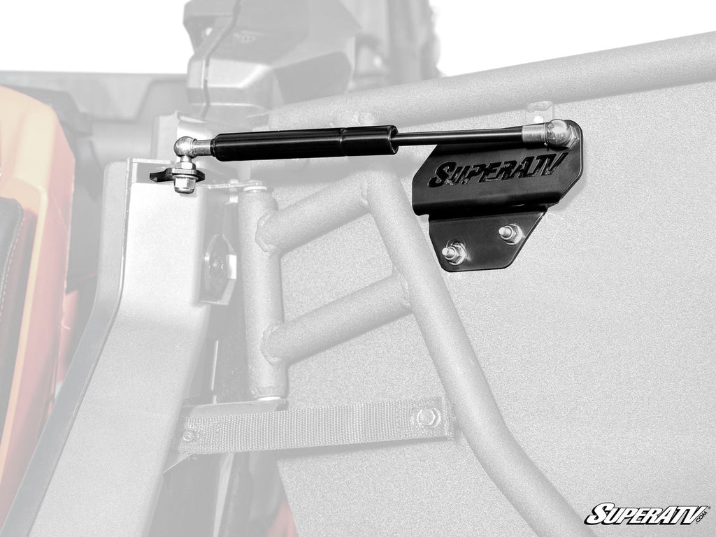 Close-up of a Can-Am Maverick X3 Gas Strut Kit for Doors by SuperATV mounted to a metal frame, showing detailed hardware and bracket, with the background and surrounding structure faded in grayscale.