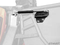 Close-up of a Can-Am Maverick X3 Gas Strut Kit for Doors by SuperATV mounted to a metal frame, showing detailed hardware and bracket, with the background and surrounding structure faded in grayscale.