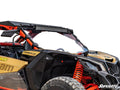 A close-up of a gold and black Can-Am Maverick X3 featuring the Full Windshield, a mounted LED light bar, orange suspension springs, rugged tires, and the SuperATV logo in the bottom right corner, all set against a white background.