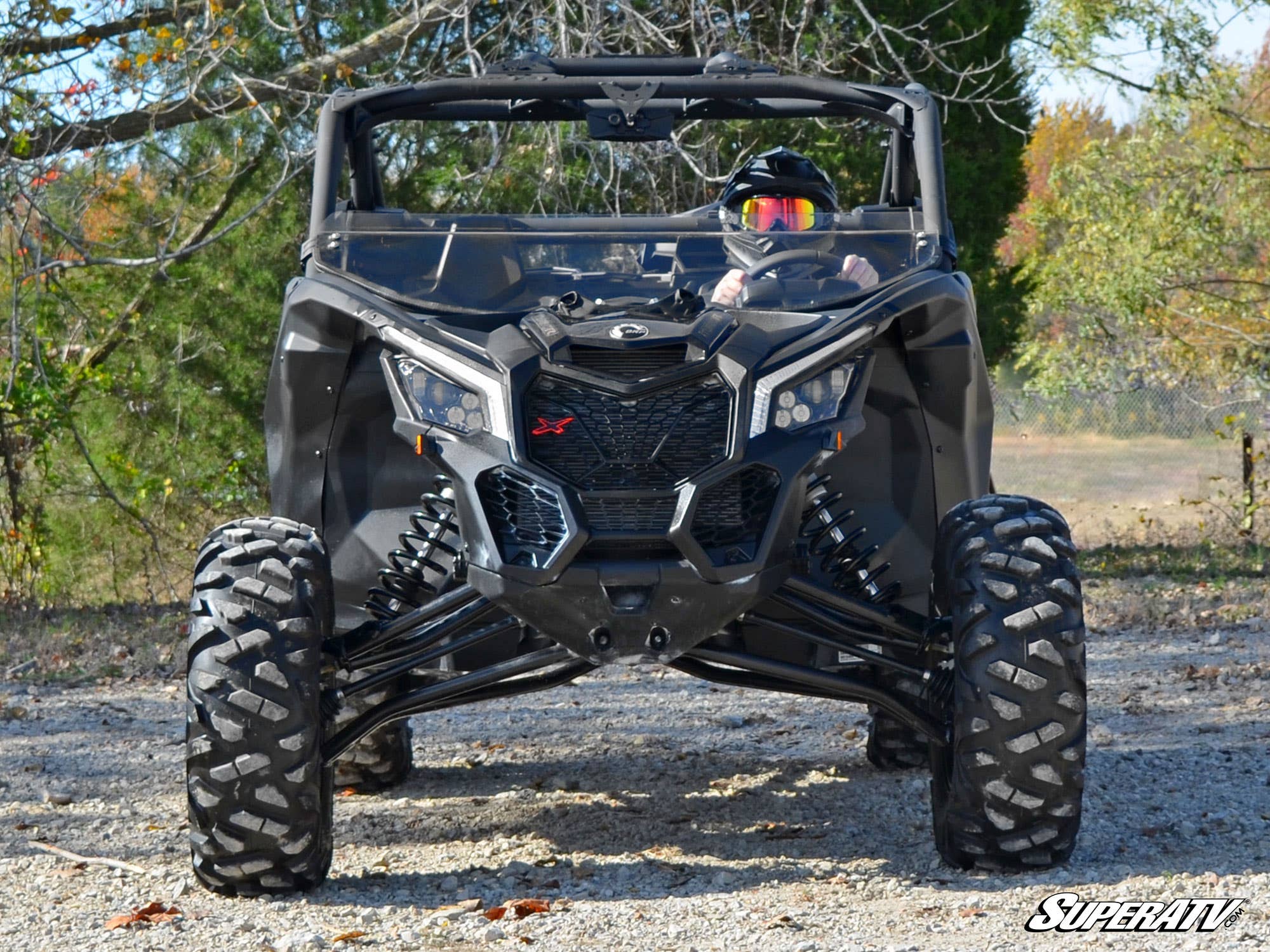 A rider in a helmet drives a black Can-Am Maverick X3 with large rugged tires along a gravel path bordered by colorful trees. The ATV features an angular front and is equipped with the Can-Am Maverick X3 Half Windshield. SuperATV logo is shown bottom righ