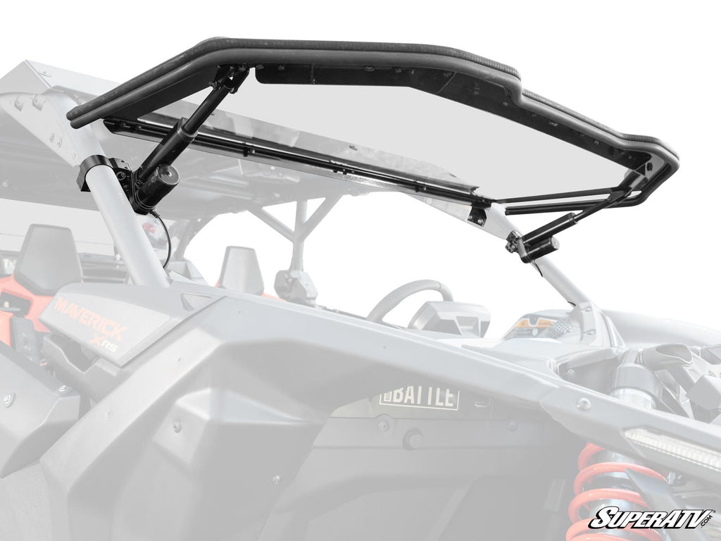 A close-up of a Can-Am Maverick X3 MaxDrive Power Flip Windshield, shown tilt-open with black metal brackets attached, reveals the UTV’s cockpit and roll cage. The SuperATV logo appears in the bottom right corner.