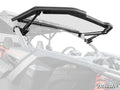A close-up of a Can-Am Maverick X3 MaxDrive Power Flip Windshield, shown tilt-open with black metal brackets attached, reveals the UTV’s cockpit and roll cage. The SuperATV logo appears in the bottom right corner.