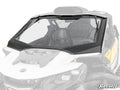A Can-Am Maverick R Glass Windshield with a black frame is installed on a white off-road vehicle, showing part of the seats. The vehicle features angular styling and displays the SuperATV logo in the lower right corner.