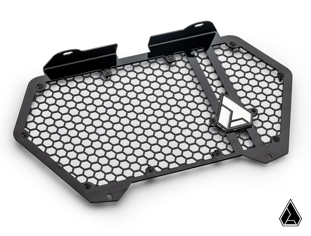 The Assault Industries Can-Am Maverick R Hellfire V2 Front Grille features a black, hexagon-patterned metal design with mounting brackets and a geometric silver emblem. It sits on a white background with a matching logo in the lower right corner, showcasi
