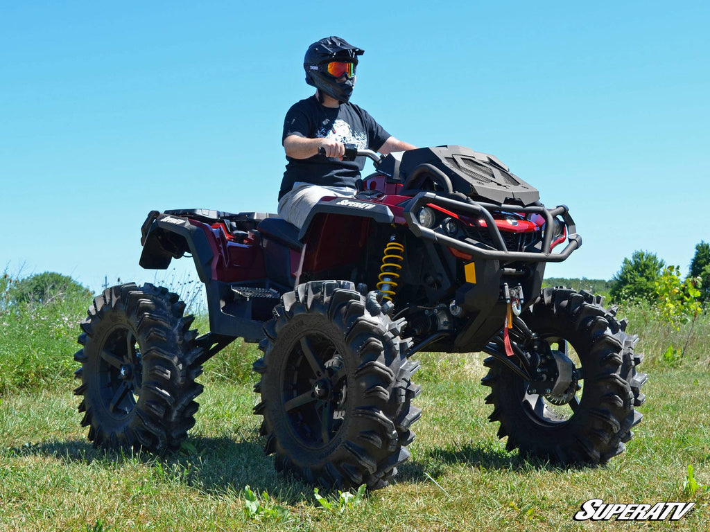Wearing a black helmet, goggles, and gloves, a rider drives a red ATV with oversized mud tires and Can-Am Renegade 4 Portal Gear Lift across grassy terrain. The ATV features yellow coil springs and gear on the front rack under a blue sky.
