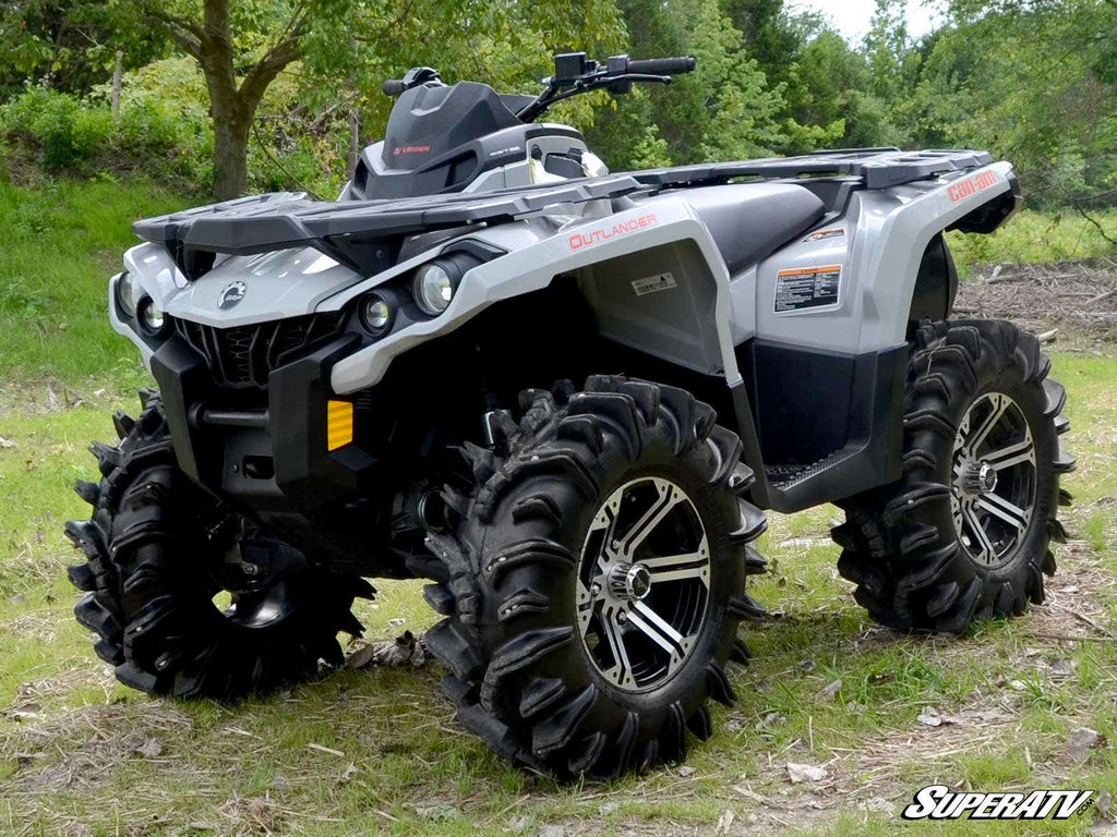 A silver Can-Am Outlander ATV with large mud tires and black alloy rims is parked on grass near trees, featuring rugged bodywork, bright headlights, and a Can-Am Outlander 2 Lift Kit logo in the lower right corner.
