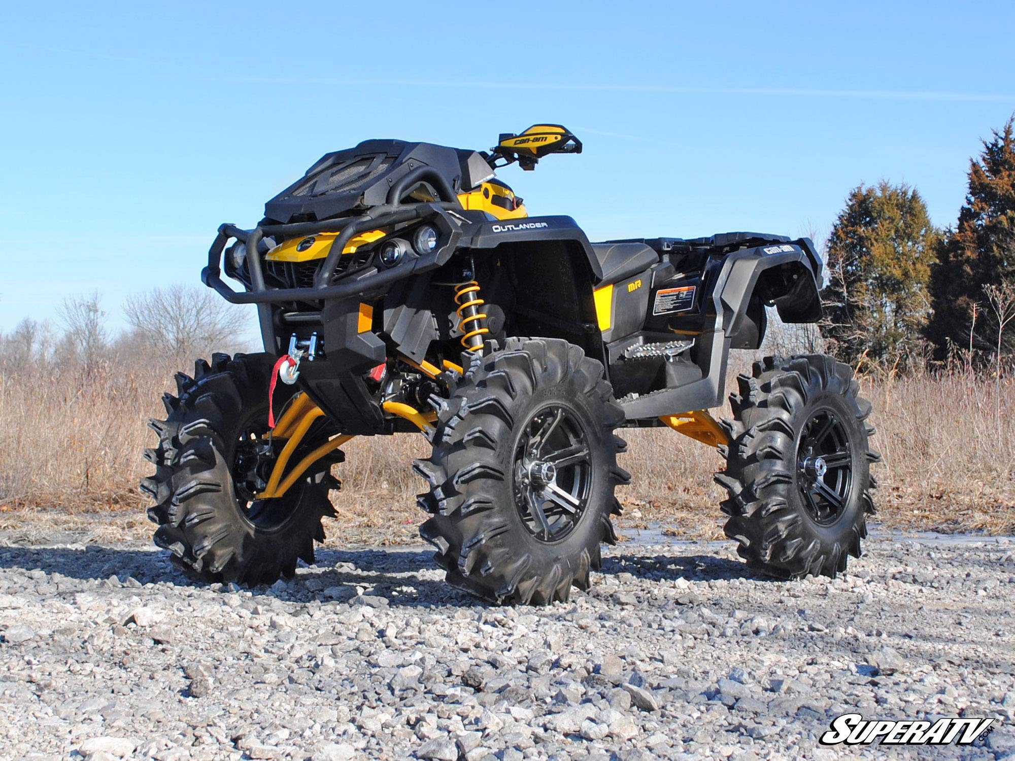 A Can-Am Renegade with a 6 Lift Kit (Gen 2), featuring bold yellow and black styling, large deep-tread tires, and elevated suspension, is parked on rocky terrain outdoors with trees and a blue sky in the background.