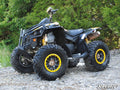 A black ATV with yellow accents and rugged tires is parked on rocky ground among green trees. It features a sturdy frame, shock absorbers, and a Can-Am Renegade (Gen 1) 2 Lift Kit. The SuperATV logo appears in the bottom right corner.