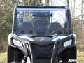 Front view of a Can-Am Maverick Sport with a black full windshield, angular headlights, two bucket seats, and the SuperATV logo. Leafless trees and grass are in the background.