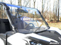 A Can-Am Maverick Trail Full Windshield is installed on a white off-road utility vehicle parked outdoors, surrounded by leafless trees and brown grass. The SuperATV logo appears in the bottom right corner.