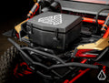 Close-up of the rear cargo area of a Can-Am Maverick X3 fitted with the Assault Industries Cooler/Cargo Box, featuring a black hard plastic design with geometric logo, secured by latches. Red shock absorbers and gold body panels surround the box.
