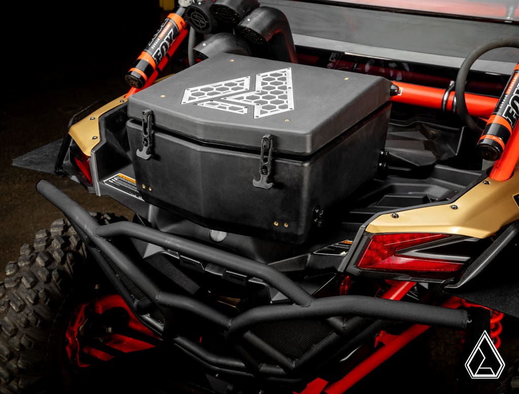 Close-up of the rear cargo area of a Can-Am Maverick X3 fitted with the Assault Industries Cooler/Cargo Box, featuring a black hard plastic design with geometric logo, secured by latches. Red shock absorbers and gold body panels surround the box.