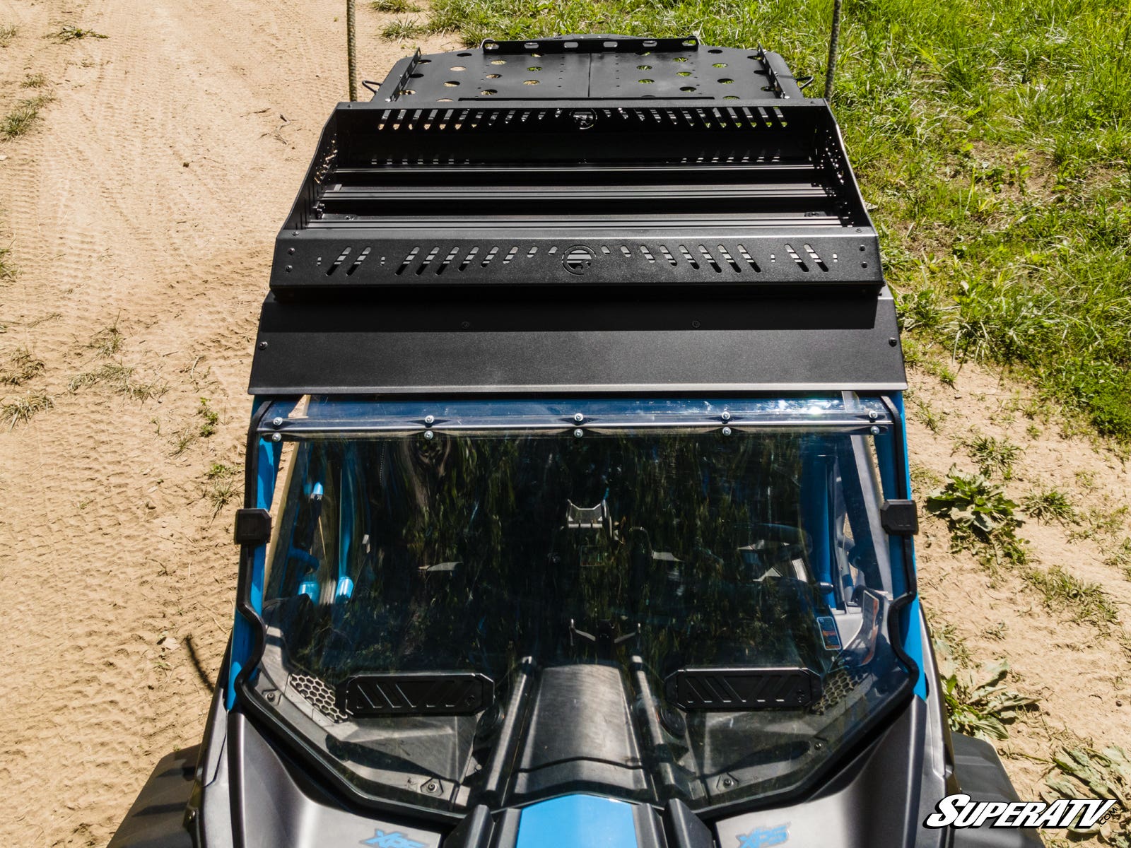 Overhead view of a blue off-road vehicle equipped with the Can-Am Maverick X3 Outfitter Sport Roof Rack, featuring a clear windshield. The vehicle is on a sandy dirt path with grass nearby. The SuperATV logo appears in the lower right corner.