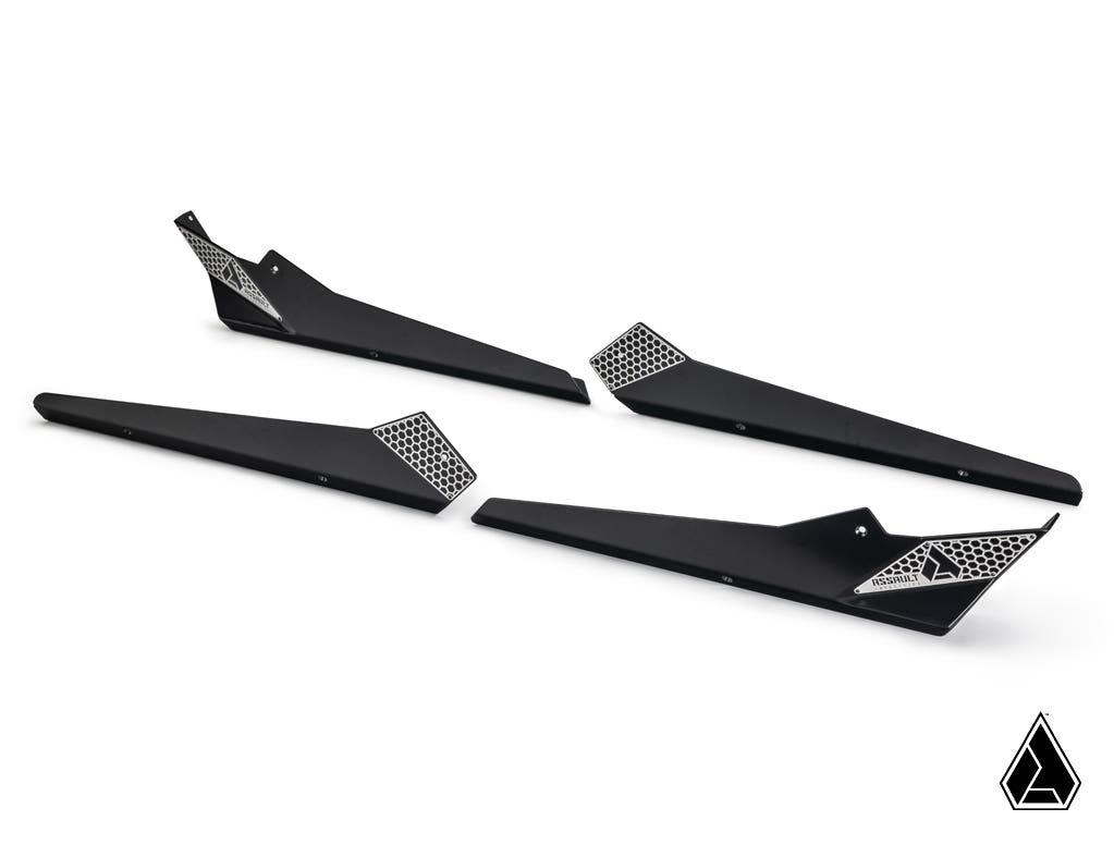 Four matte black Assault Industries Can-Am Maverick X3 MAX Rock Sliders with a geometric honeycomb pattern and logo on one end are arranged diagonally on a white background. A small triangular black-and-white logo appears in the lower right corner.