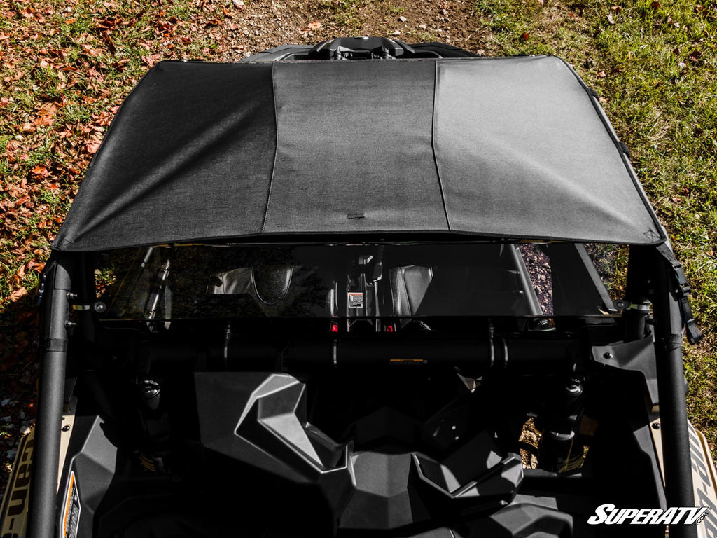Overhead view of a black UTV with the Can-Am Maverick X3 Primal Soft Top Roof parked on grass and fallen leaves, showing its angular dashboard and roll cage. Sunlight casts shadows across the roof, with the SuperATV logo in the lower right corner.